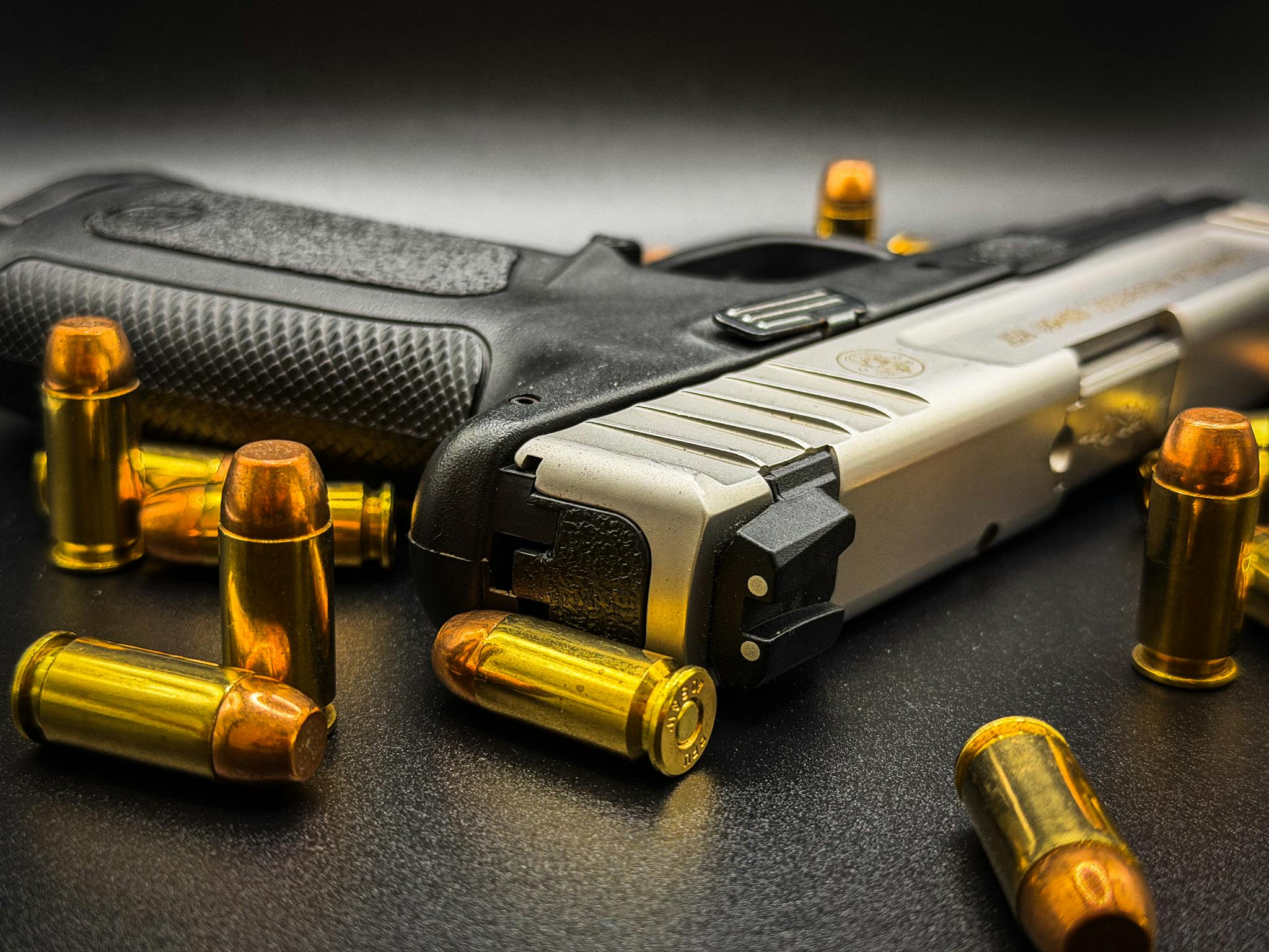 Detailed image of a handgun surrounded by bullets on a black surface, showcasing its metal finish and texture.